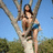 A pixelated image shows a person with long dark hair standing outdoors under a clear blue sky, holding up two tree branches against green foliage.