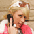 A pixelated image of a person with light brown hair holding a black phone to their ear, wearing a pink top against a beige background.