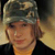 Static 50x50 pixel icon showing a person wearing a camouflage-patterned cap with visible texture details, brown hair peeking out from under the cap, and a dark jacket over an orange shirt against a gradient background.