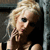 Pixelated image of a person with light-colored hair styled in an updo, wearing dark clothing and a necklace, positioned against a dark background with a vertical light-colored structure on the right.