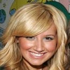 A static close-up of a person with long blonde hair and a side-swept fringe, smiling with visible teeth against a blurred background with blue and yellow shapes.