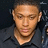Pixelated close-up of a person with short light brown hair wearing dark clothing against a dark background.