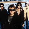 Static image of four people standing together in a group, likely band members, with varied clothing and sunglasses against an outdoor background.