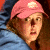 Pixelated image of a person wearing a pink headscarf with a white circular mark, brown hair, and a blue garment, set against a dark background.