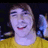 Pixelated static image of a person with light skin tone wearing a yellow top, smiling with visible teeth, against a dark background with blue and yellow elements.