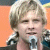 Pixelated image of a person with light brown hair holding a microphone against an orange background with blue and yellow elements.