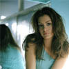 A woman with long wavy brown hair wearing a light blue sleeveless top, positioned slightly off-center against a blurred background with light blue and white tones.