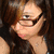Pixelated close-up of a person with dark hair and glasses, wearing a light-colored top against a dark background.