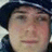 Static pixelated image of a person wearing dark headwear with light trim, visible facial features including eyes and mouth, and blurred background elements.