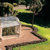 A pixelated static image shows a small house with a light gray roof and brown walls, situated on a grassy area with a winding path leading to it under a dark tree canopy.