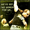 Pixelated image of a person in a black shirt with white text, holding a microphone, with text overlay reading "WE'VE GOT TWO WORDS FOR YA..." on a dark background.