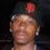 A person wearing a dark cap with a red emblem, smiling with eyes closed, against a dark background.