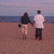 Two figures walk hand-in-hand on a beach toward the ocean under a pale sky.