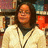 Pixelated image of a person with long dark hair wearing light-colored clothing and multiple necklaces against a blurred background.