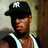 Pixelated image of a person wearing a black cap with a white emblem, dark shirt, and white straps over the shoulders, standing against a textured wall and wooden structure.