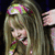 A pixelated image of a person with long light brown hair wearing a colorful flower crown, holding a small object near their face against a dark background.