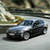 A pixelated image of a silver sedan with dark tinted windows driving on a road, background shows blurred greenery and sky.