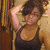 A person in a black tank top adjusts their hair with both hands against a background of vertical striped fabric.