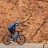 A pixelated figure in blue and dark clothing rides a bicycle against a brown textured background with a light strip at the bottom.