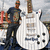 A person stands beside a white electric guitar in an outdoor setting with blurred background elements.