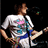 A person in a white t-shirt with blue and pink graphics plays a light-colored electric guitar against a dark background.