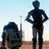 Pixelated static image of two humanoid figures in dark blue and black silhouettes against a light sky background with brown ground, one standing tall on the right and one crouching or kneeling on the left holding an object.