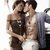 Two people in a close embrace, one shirtless with a gray jacket draped over the shoulder, the other in a dark strapless top, both with brown hair and white pants, in a static pose.