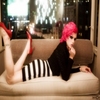 A person with bright pink hair lies on a beige couch, wearing a black top and black-and-white striped shorts, with red high-heeled shoes visible.