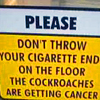 A static sign with a white top section and yellow bottom section displaying bold text about not throwing cigarette ends due to cockroaches getting cancer.