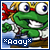 Green cartoon frog with large white eyes and blue pupils, wearing white shirt and red headband, smiling with visible teeth, standing on brown platform with blue sky background.