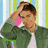 Pixelated image of a person with brown hair wearing a green jacket over white shirt, holding pink object near face against striped background.