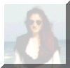 A woman with long brown hair and sunglasses stands against a beach background, wearing a white top under a dark vest.