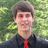 Pixelated portrait of a person with short brown hair wearing dark clothing and red tie against green foliage background.