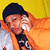 A person in an orange hoodie and dark cap covers their mouth with a white cloth against a yellow background.