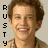 Pixelated portrait of a smiling person with brown hair, vertical black text 'RUSTY' on left side, light beige background.