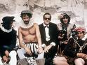 Static image of five people in historical costumes sitting together against a stone background.