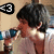 Pixelated image of a person with dark hair wearing a light-colored shirt, holding a blue object near their face, with a black and white '63' symbol in the top-left corner.