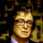 Pixelated static image of a character with light brown hair and glasses against dark background with golden decorative elements.