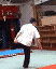 A person in a white shirt and dark pants performs a sequence of acrobatic movements on a blue mat with red borders, against a backdrop of stacked boxes.