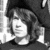 Black-and-white pixelated image of a person with shoulder-length hair wearing a dark top, positioned against a light background with vertical lines.