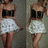 A pixelated static image shows a person in a white dress with black straps and brown hair, standing against a gray background.