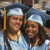 Two people wearing graduation caps and gowns stand side-by-side, smiling at the camera with a blurred background.