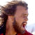 Pixelated static image of a person's face and upper body in profile view with visible facial hair, dark hair, and red clothing against a light background.