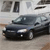 A dark sedan with visible headlights and side mirrors is positioned on a light-colored surface, with a blurred background showing indistinct shapes and colors.