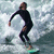 Pixelated image of a person in a black wetsuit surfing on a wave, arms outstretched, legs bent, feet on a light green surfboard.