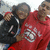 Two people in red hoodies with white text pose together, one holding a black steering wheel prop against a light background.