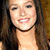 A pixelated close-up of a person with long dark hair, light skin tone, and visible teeth in a smiling expression against a blurred background.
