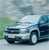 Static pixelated image of a light-colored sedan with dark blue accents, positioned on a road with green and gray background elements.