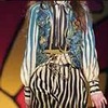 A static icon shows a person with long light-colored hair wearing a striped top with a patterned section above the waist, set against a blurred background with red and yellow tones.