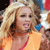 A pixelated figure with long blonde hair wears an orange top with a red strap, holding a blue object near their face against a blurred background.